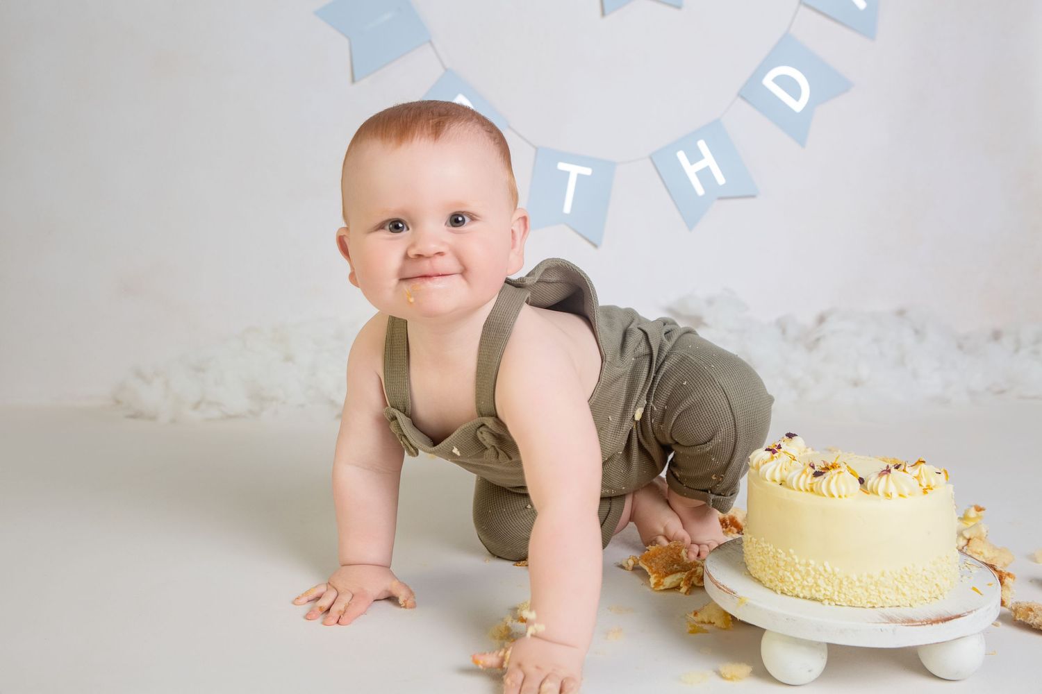 Baby boy in olive romper crawls toward a smashed yellow birthday cake under a blue BIRTHDAY banner.