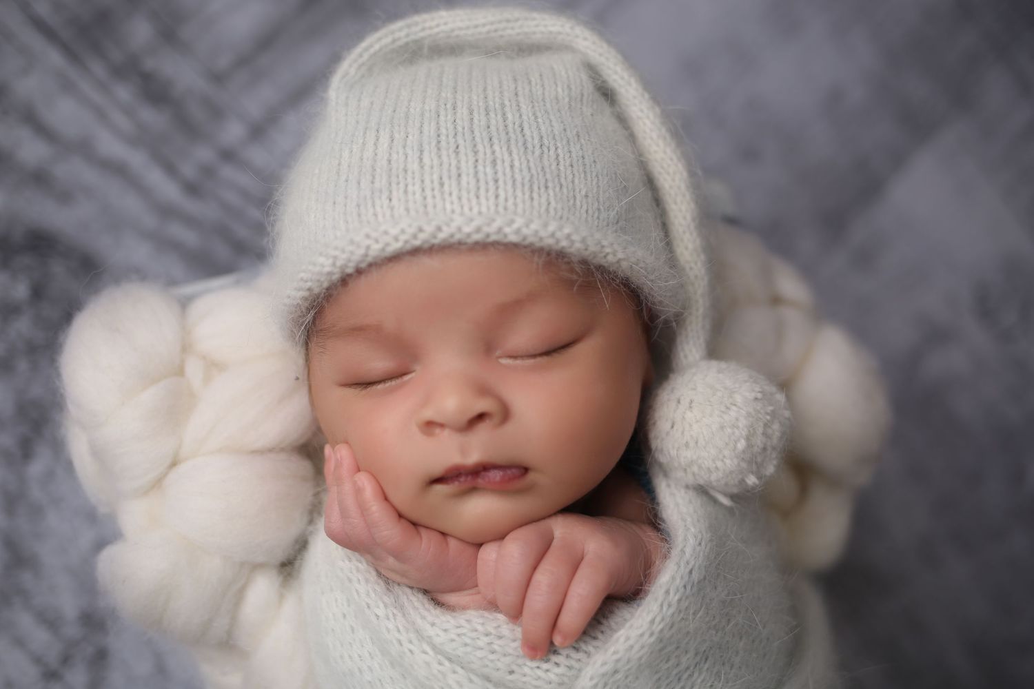 Peaceful newborn baby sleeping in white knit wrap and sleepy hat with pom-pom on gray background.