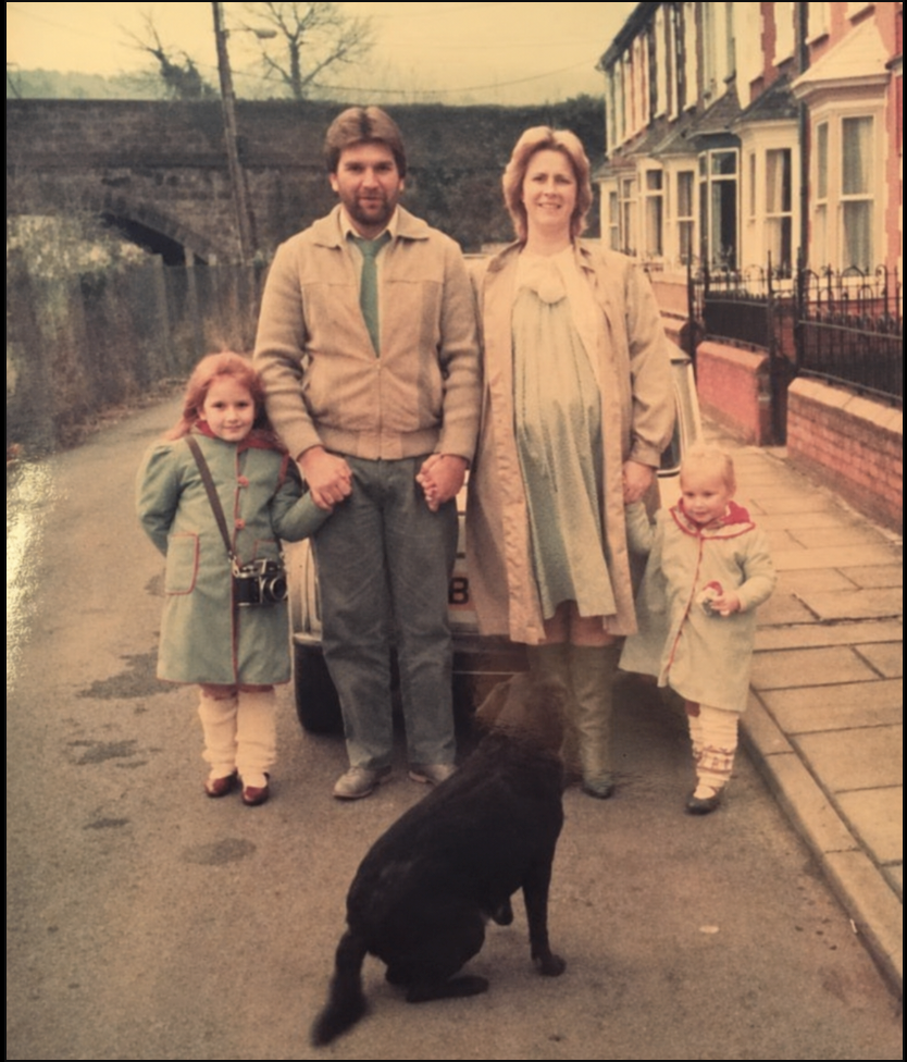 Vintage family photo of parents with two young children and black dog on a residential street.