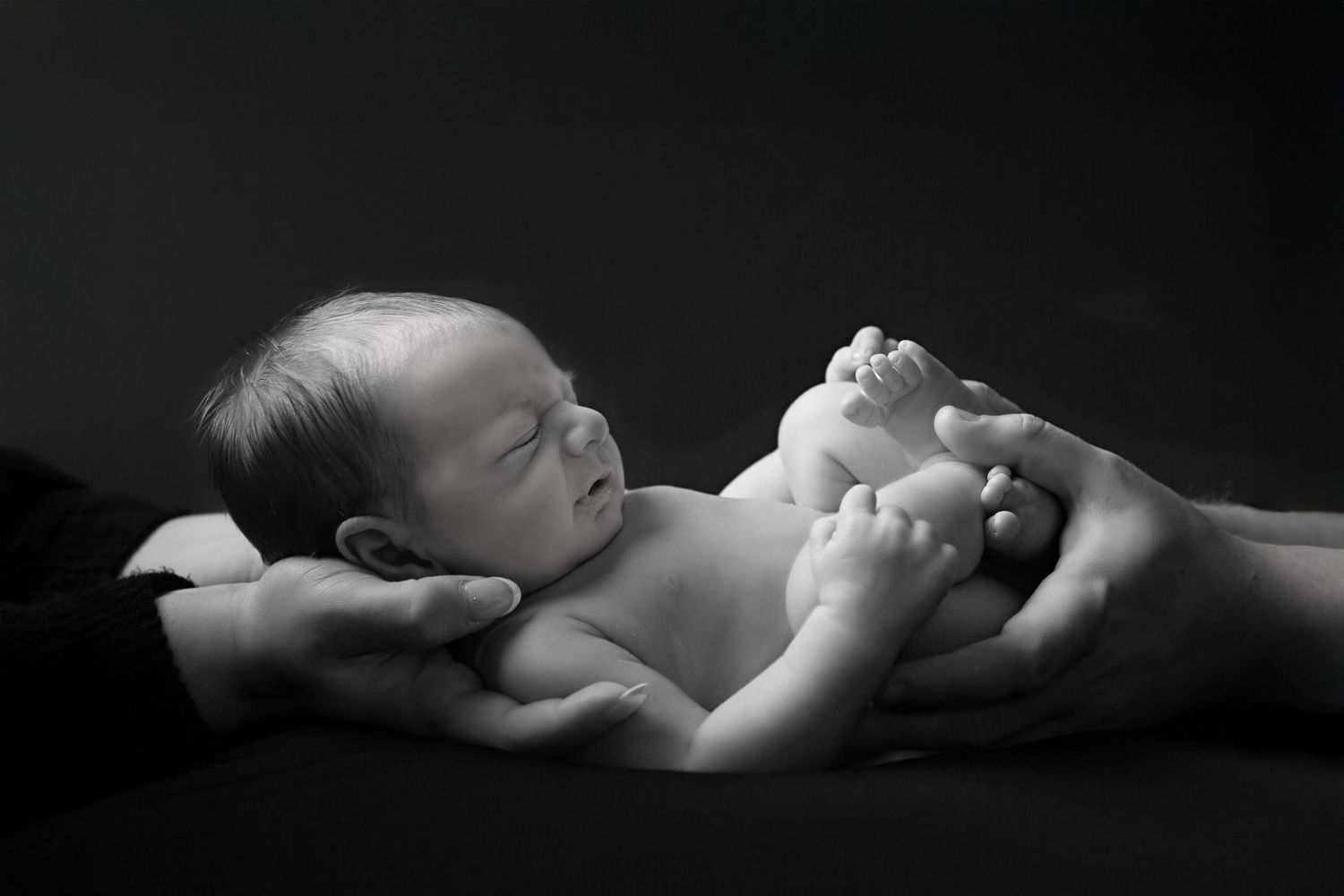 Black and white newborn photography of a sleeping baby cradled gently in adult hands against dark background.