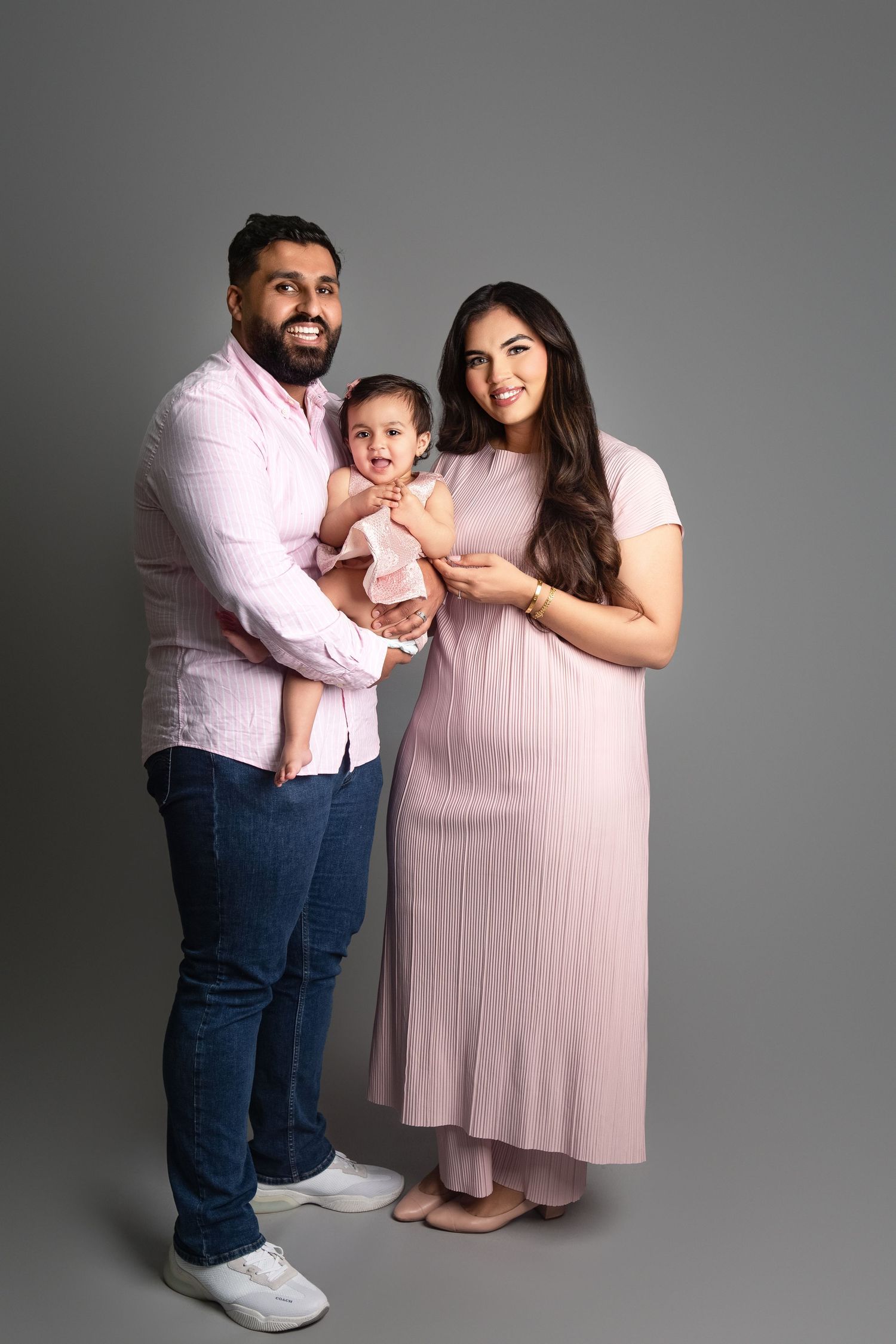 A family of three poses together, parents in pink outfits holding their baby girl against a gray backdrop.