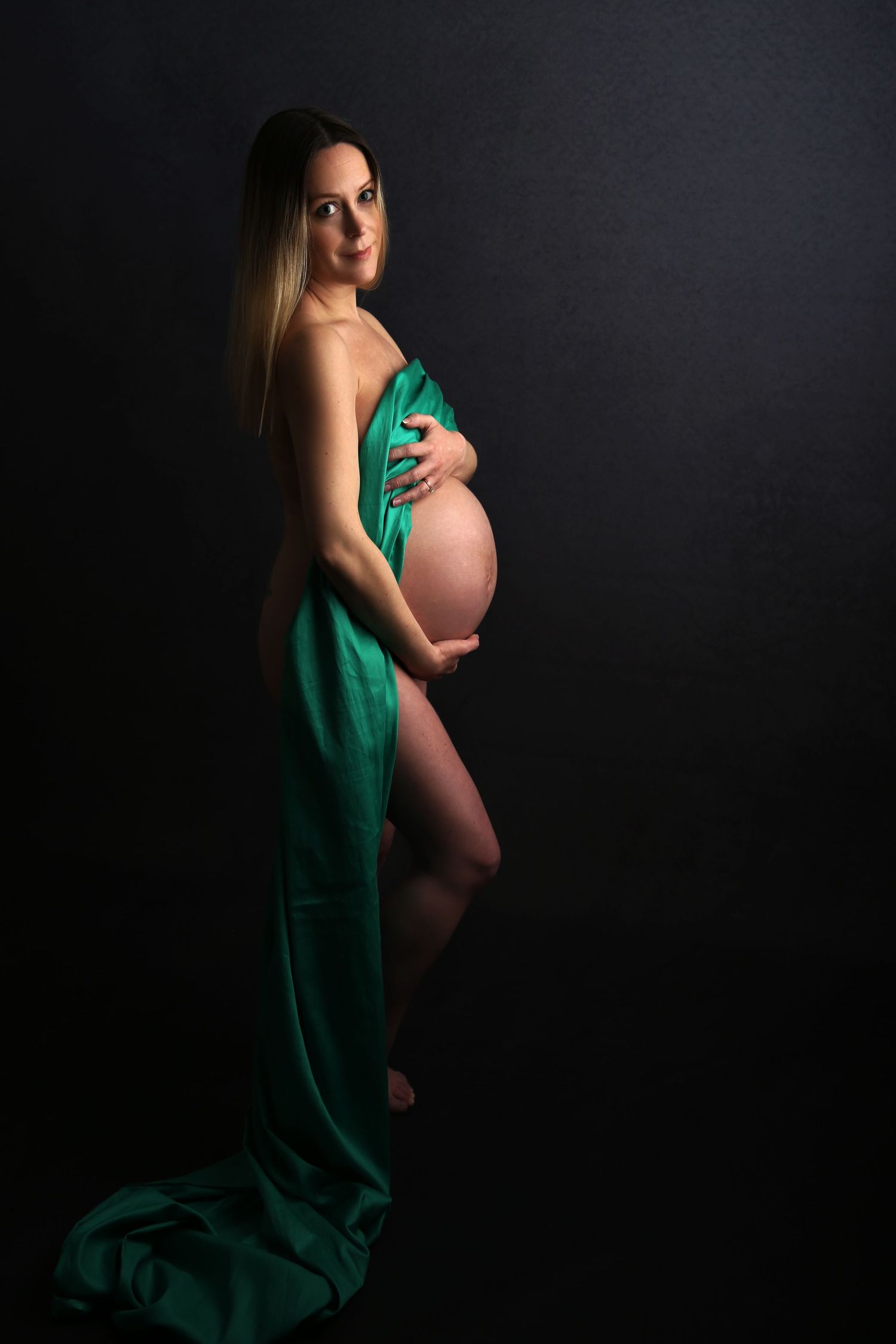 Pregnant woman draped in green silk fabric posing against dark background, cradling her baby bump in a maternity photo.