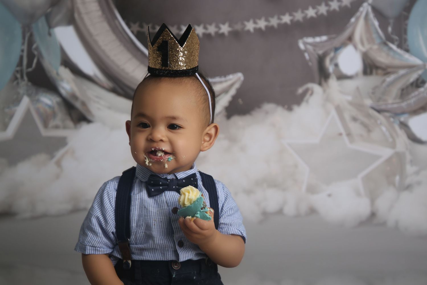Adorable baby boy in crown headband and bow tie suspenders celebrating first birthday with cake smash against star backdrop.
