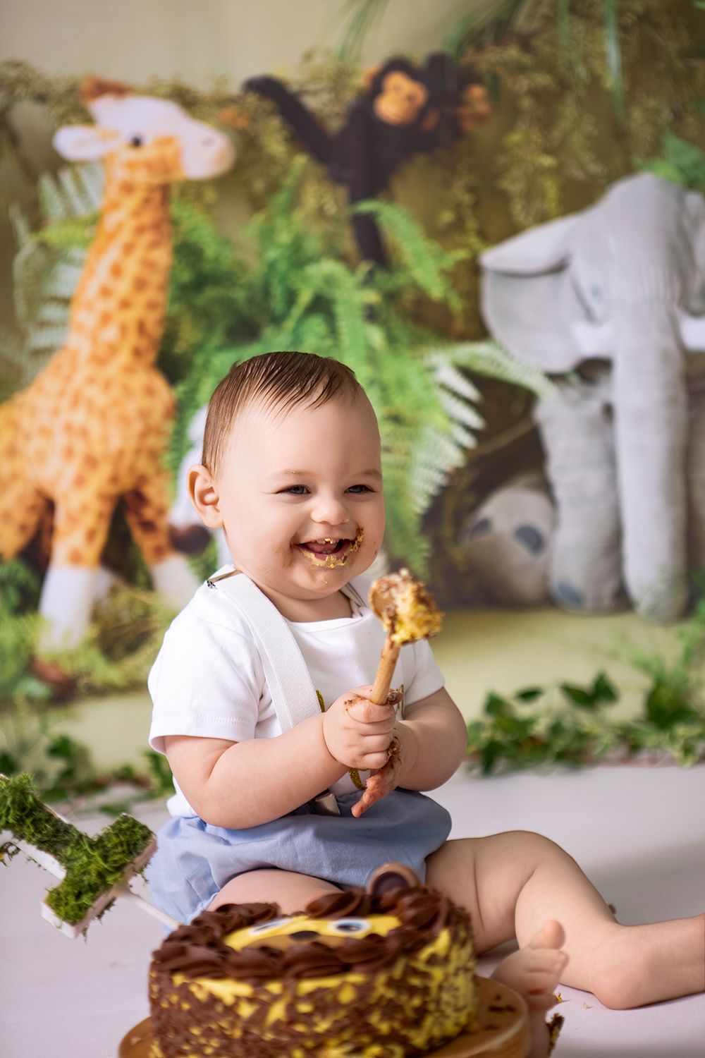 Smiling baby boy celebrating first birthday with jungle safari cake smash, chocolate cake and giraffe backdrop.