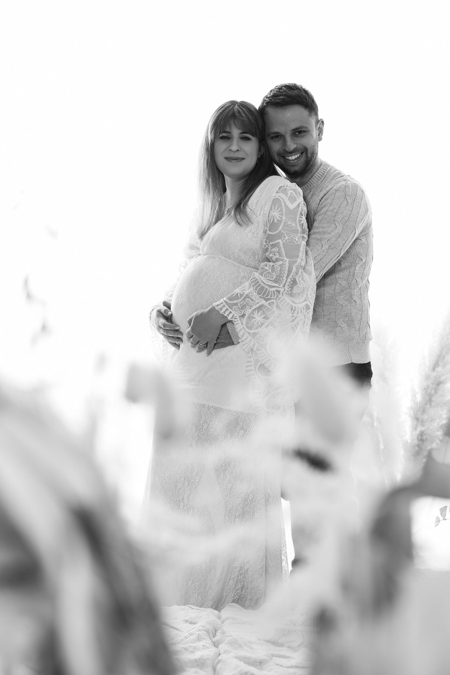 Black and white maternity photo of smiling couple embracing outdoors among tall grasses.