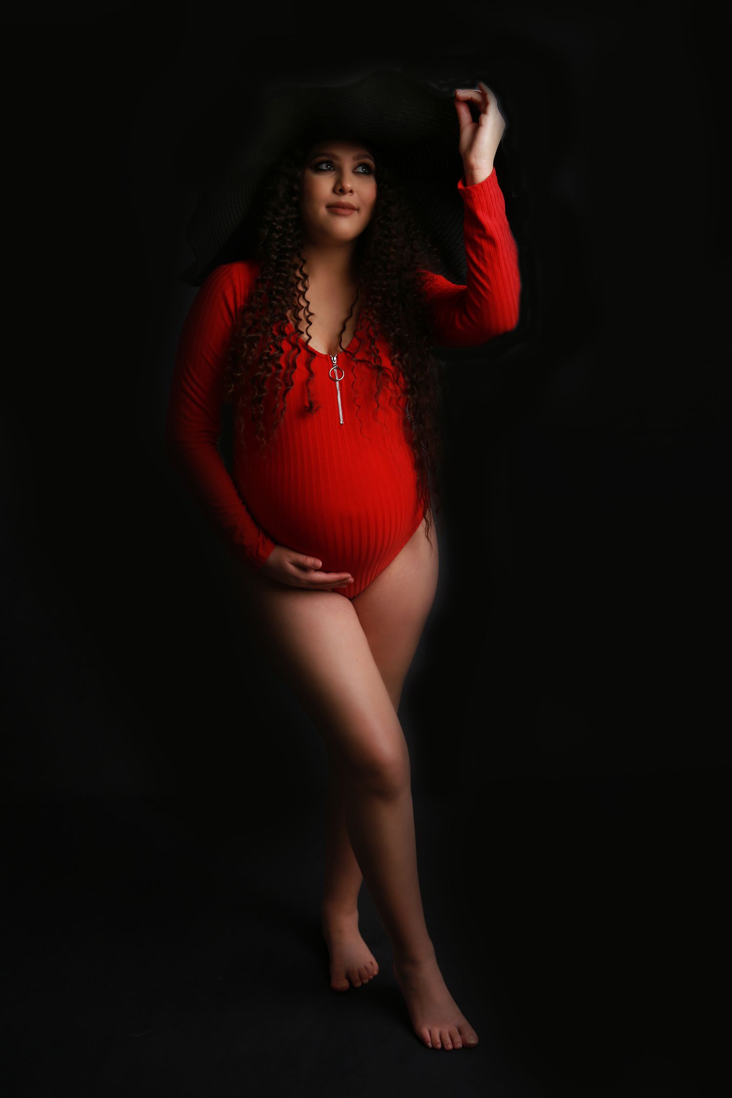 Pregnant woman with curly hair in red bodysuit and black hat in studio portrait.