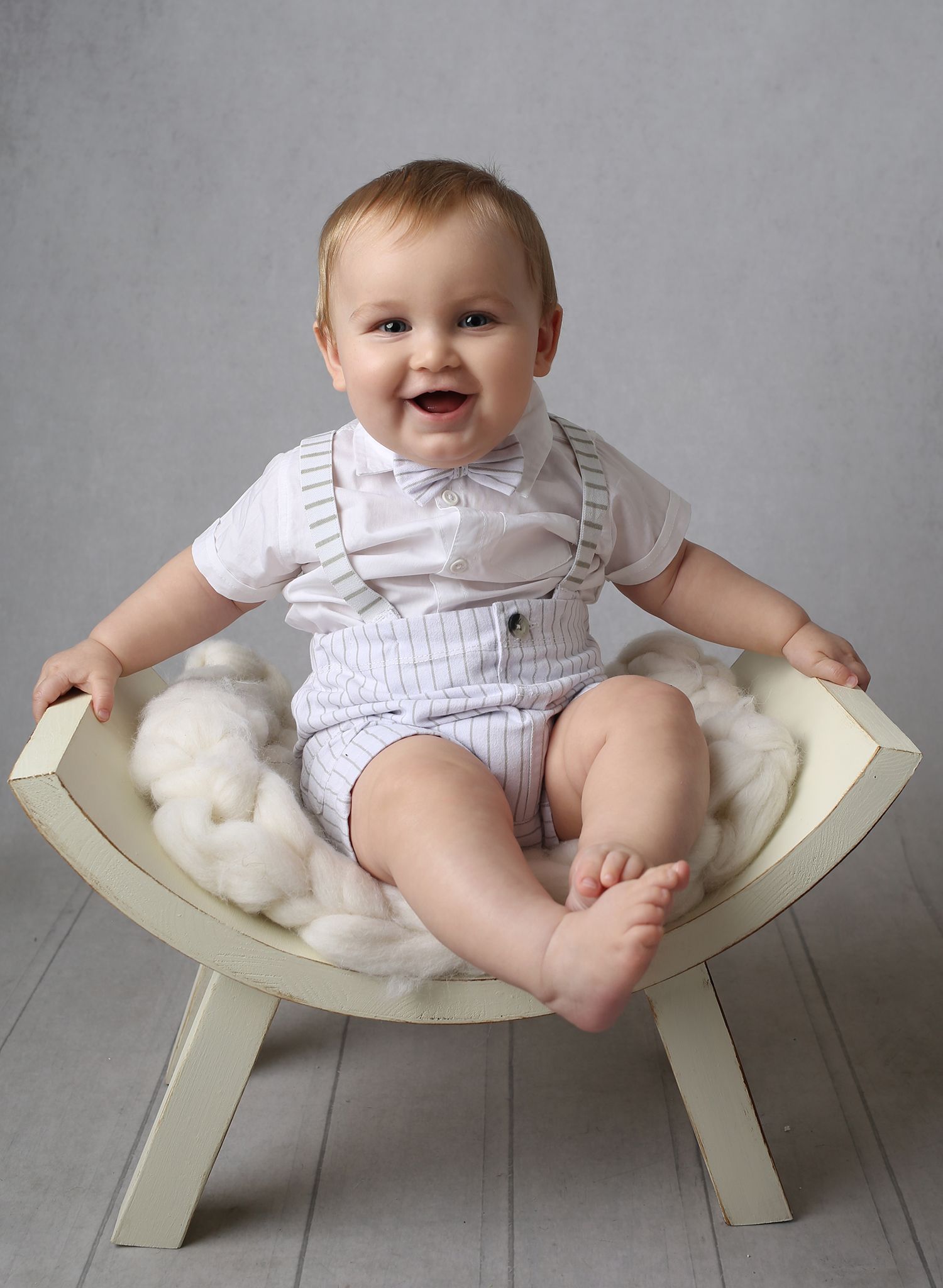 Smiling baby boy in white outfit with bow tie sits on a cushioned wooden chair for a professional portrait photo.