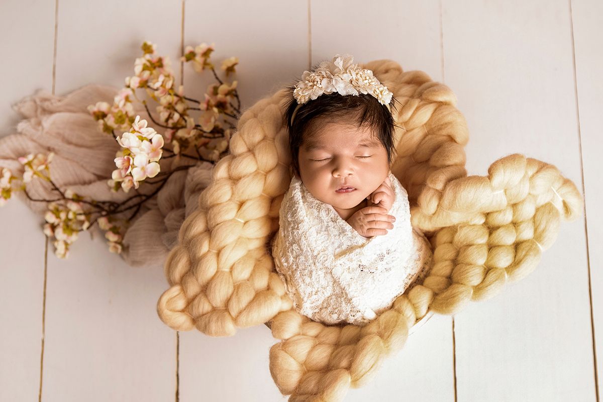 Newborn baby sleeping in chunky knit bowl wrap with lace swaddle and floral crown on white wood floor.