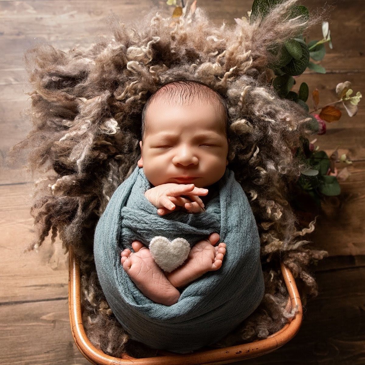 Newborn baby swaddled in teal wrap with a heart stone, nestled in fluffy brown fibers in a wooden bowl.