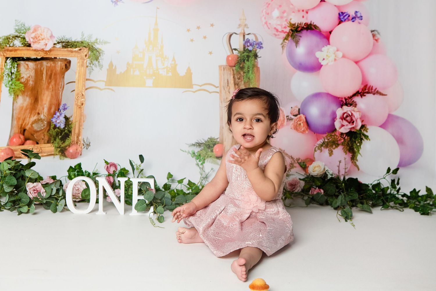 Baby girl in pink dress sits before colorful balloon arch and ONE sign at her first birthday cake smash photoshoot.