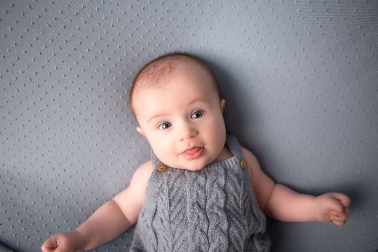 Adorable baby in gray knit romper lying on gray blanket, looking up with big eyes.