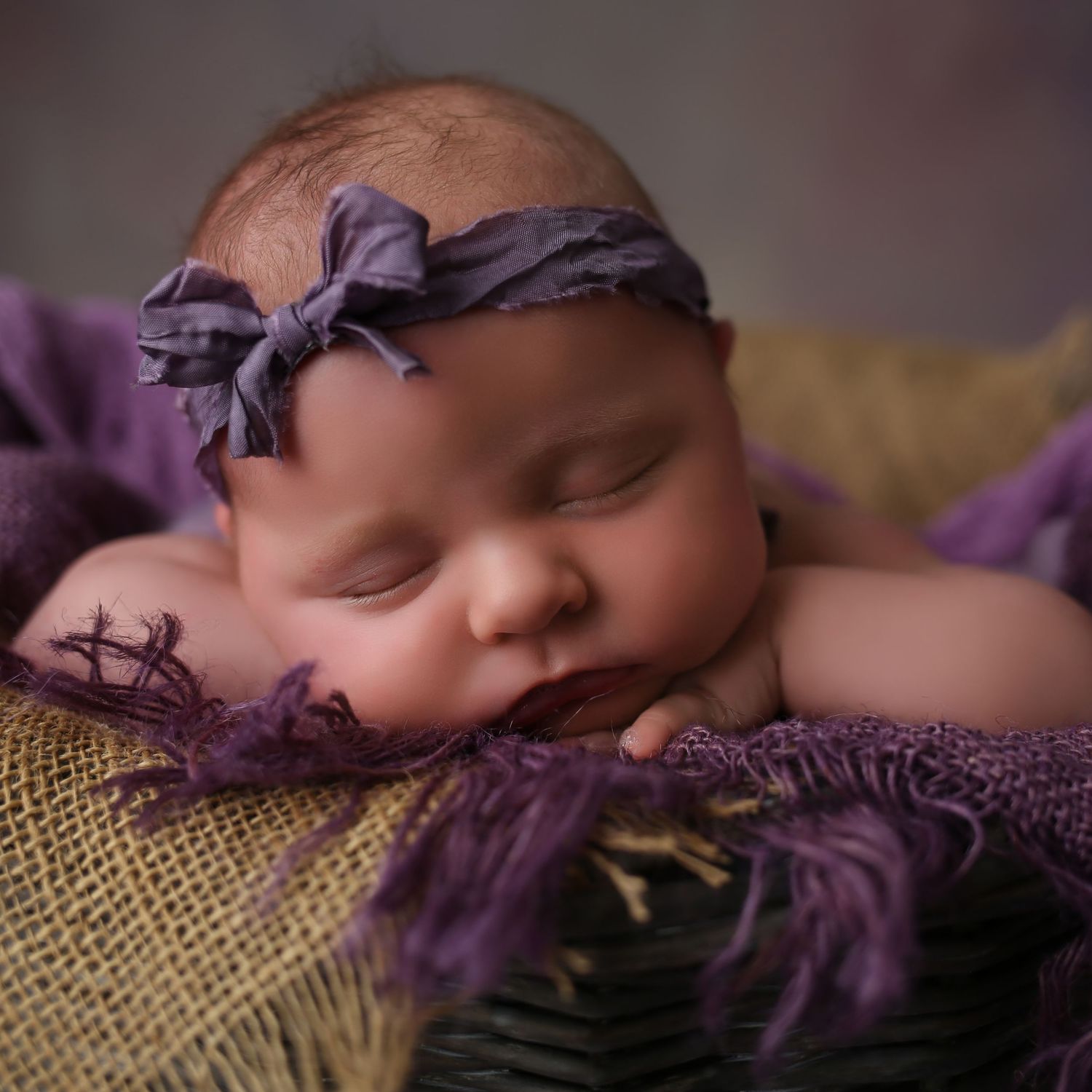 Sleeping newborn baby girl wearing a purple bow headband, nestled in a basket with purple fabric wrap.