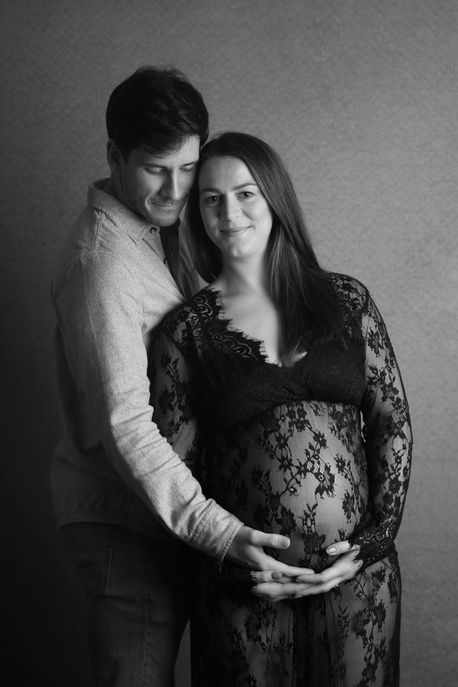 Black and white maternity photo of a couple tenderly cradling a pregnant belly in a lace dress.