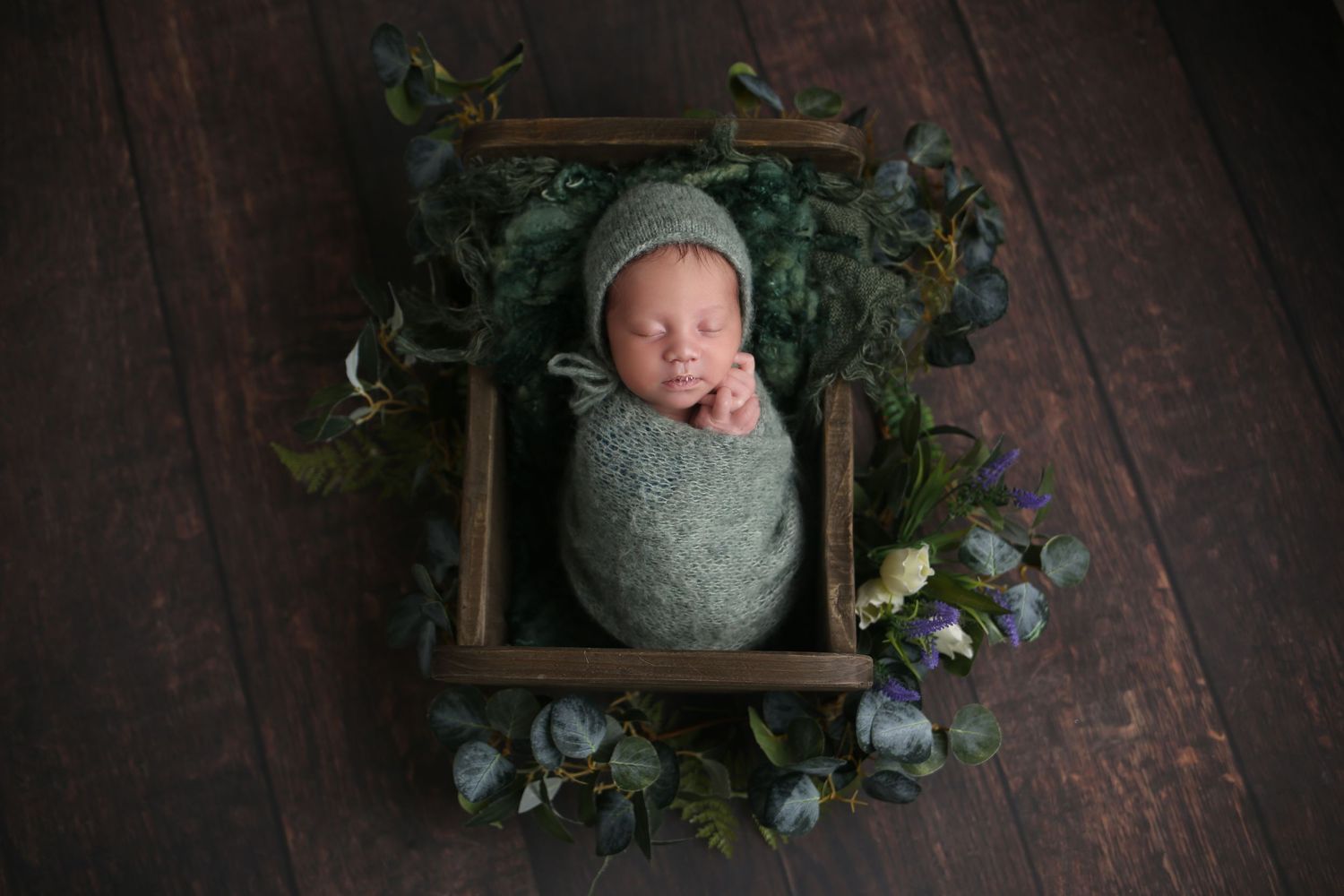 Newborn baby wrapped in green swaddle sleeping in a wooden mini crib surrounded by flowers.