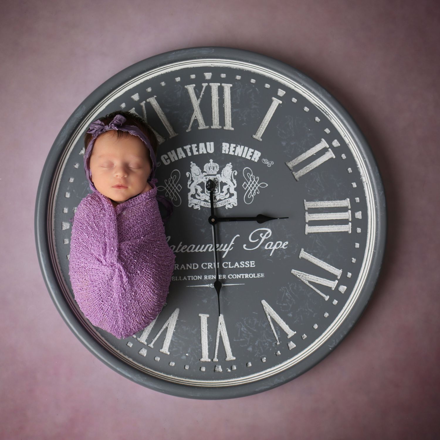 Newborn baby wrapped in purple swaddle sleeping on a large vintage Chateau Remier decorative wall clock.