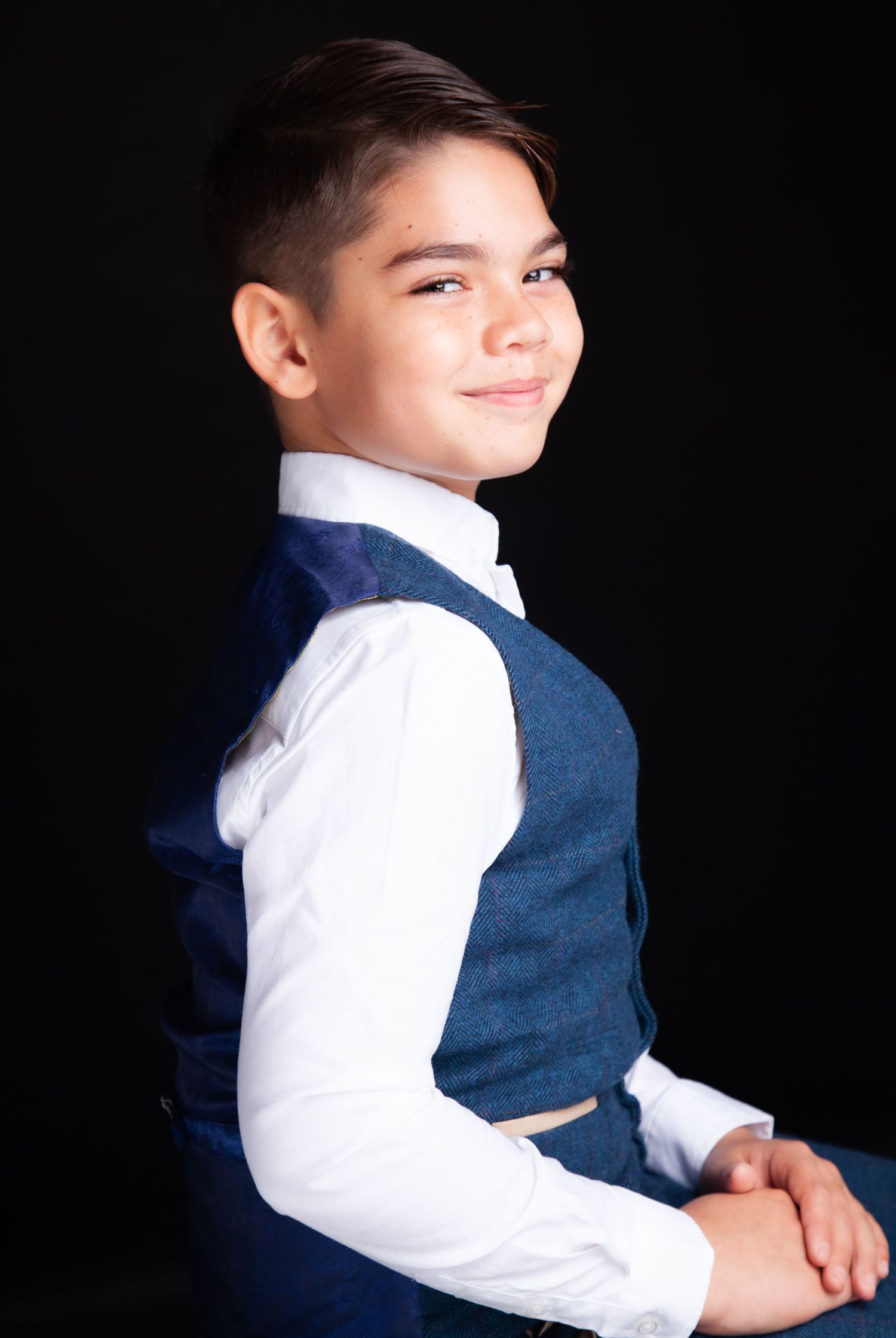 A young boy in a white dress shirt and blue vest smiles confidently against a dark background.