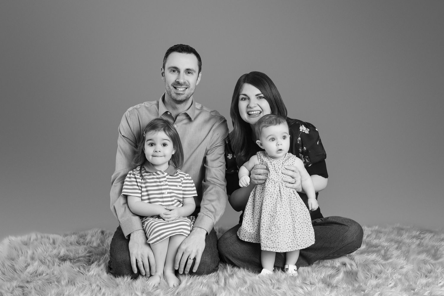 Black and white family portrait of parents with two young daughters sitting on a fluffy rug against a gray backdrop.