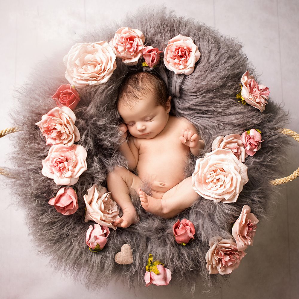 Newborn baby sleeping peacefully on gray fur wreath adorned with pink roses and heart decorations.