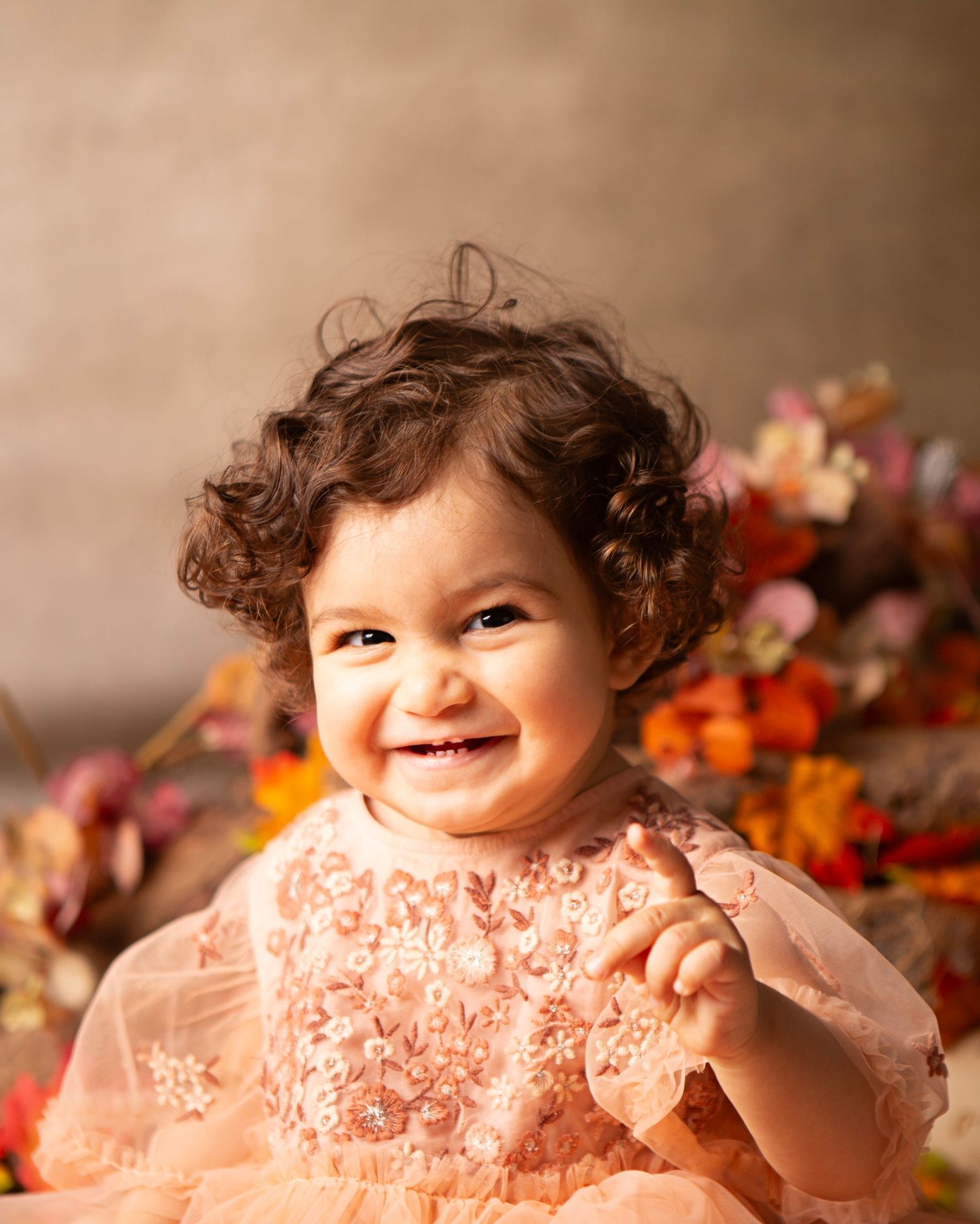 Smiling toddler girl with curly hair wearing a pink floral lace dress, surrounded by colorful flowers in a studio portrait.