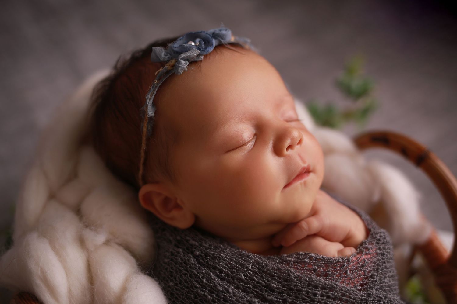 Peaceful newborn baby sleeping in a basket wrapped in gray knit, wearing a delicate blue floral headband.