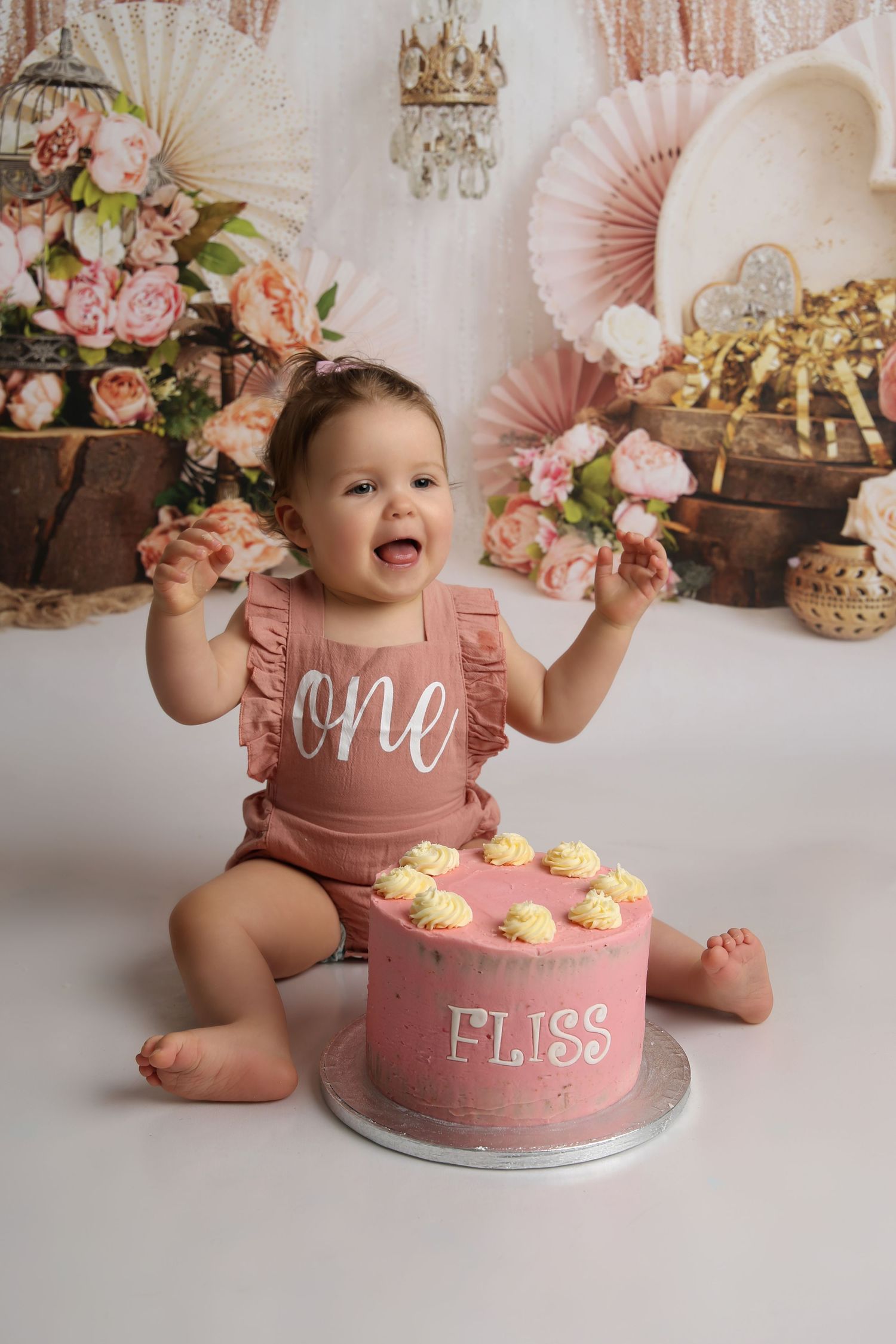Baby girl in pink romper smashing her first birthday cake labeled Fliss, surrounded by floral decorations.