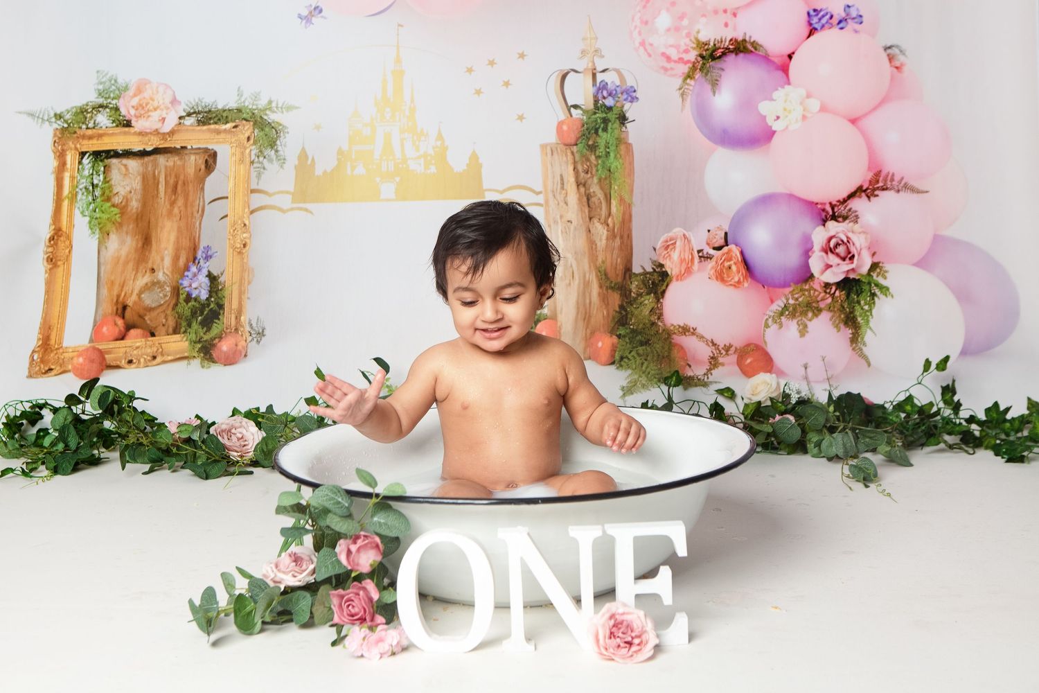 Baby sitting in a white tub with ONE sign, pink balloons, flowers, and a castle backdrop for a first birthday.