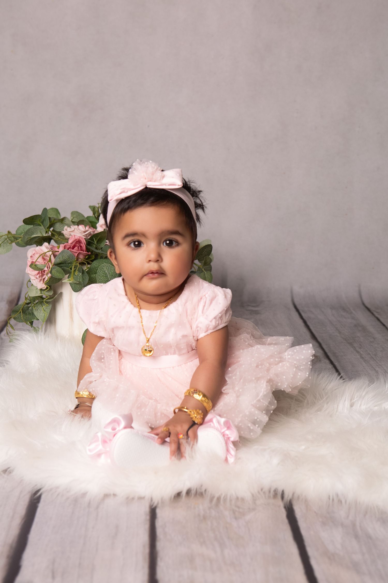 Adorable baby girl in pink tutu dress and gold jewelry sits on fluffy white rug with floral backdrop.