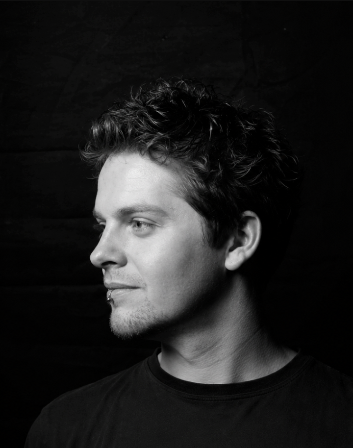 Black and white profile portrait of a young man with curly hair and a lip piercing against dark background.