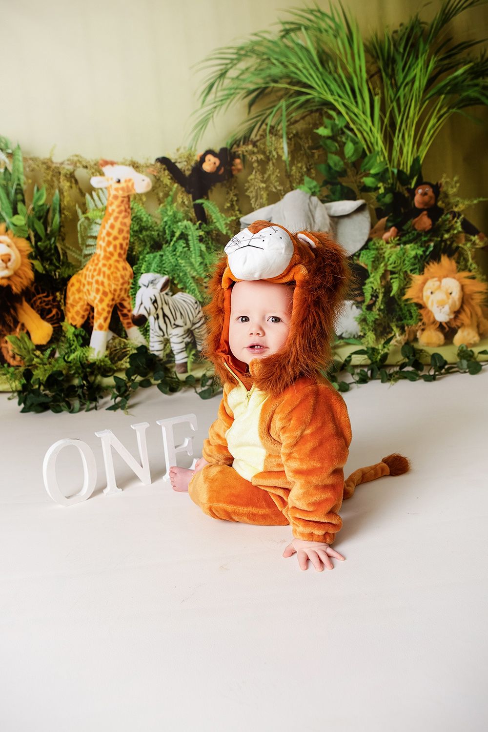 Baby in lion costume sits before jungle backdrop with stuffed animals and ONE sign at first birthday cake smash.