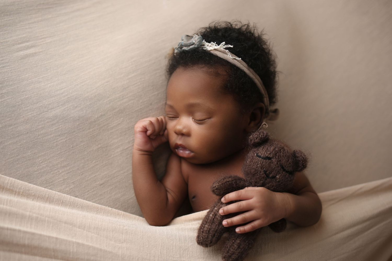 Sleeping newborn baby girl with floral headband cuddling a brown knit teddy bear on a soft beige backdrop.