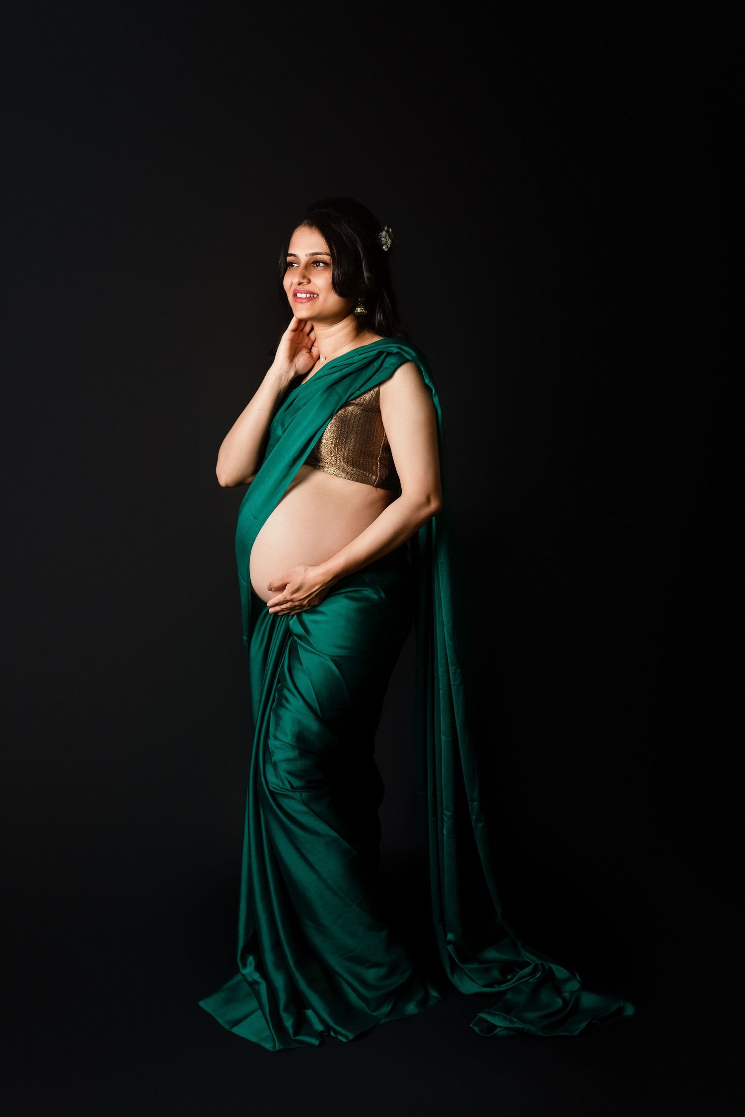 Pregnant woman in green silk saree smiling softly, cradling baby bump against dark studio background.