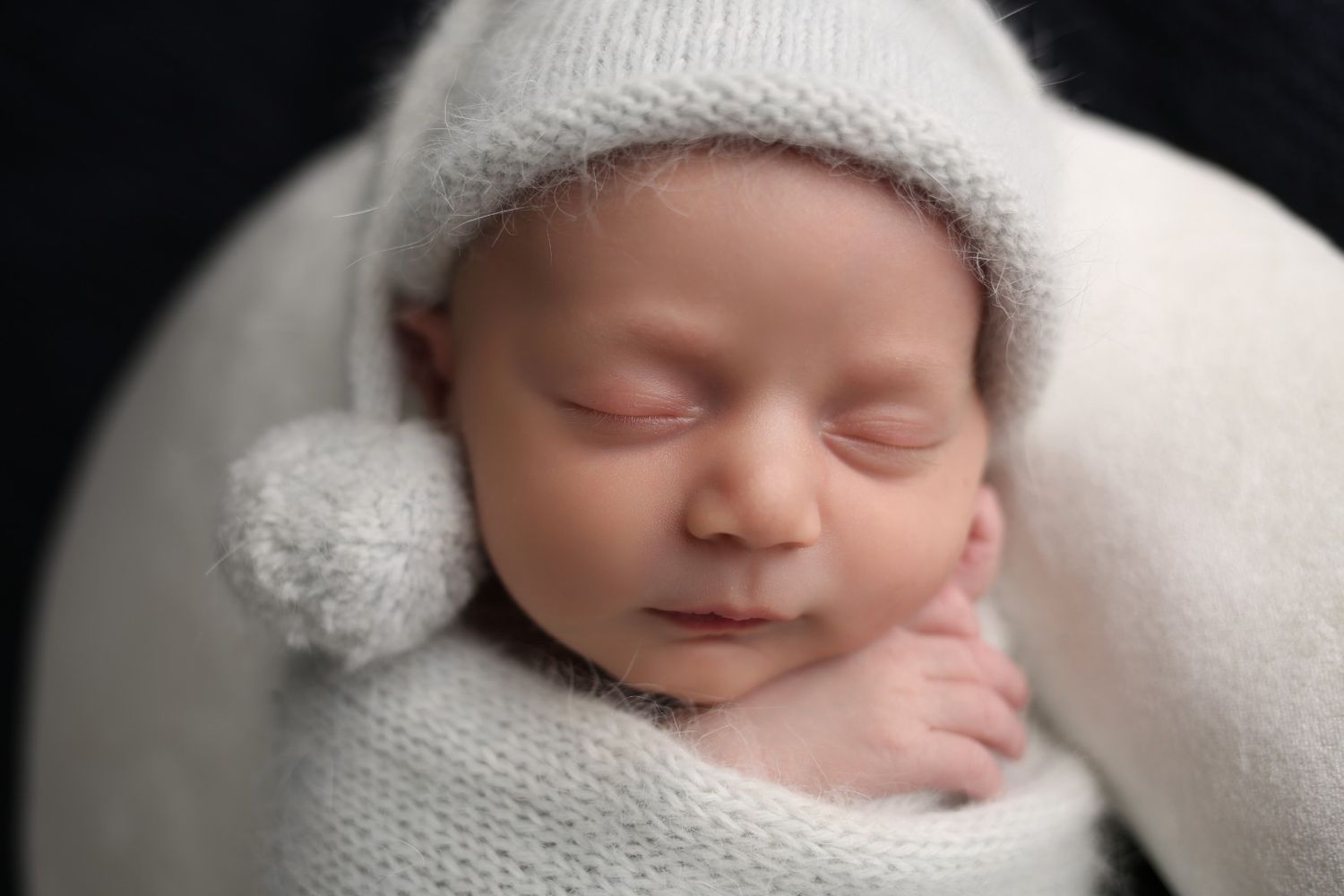 Peaceful newborn baby sleeping wrapped in white knit blanket and hat with pom-pom, professional newborn photography.