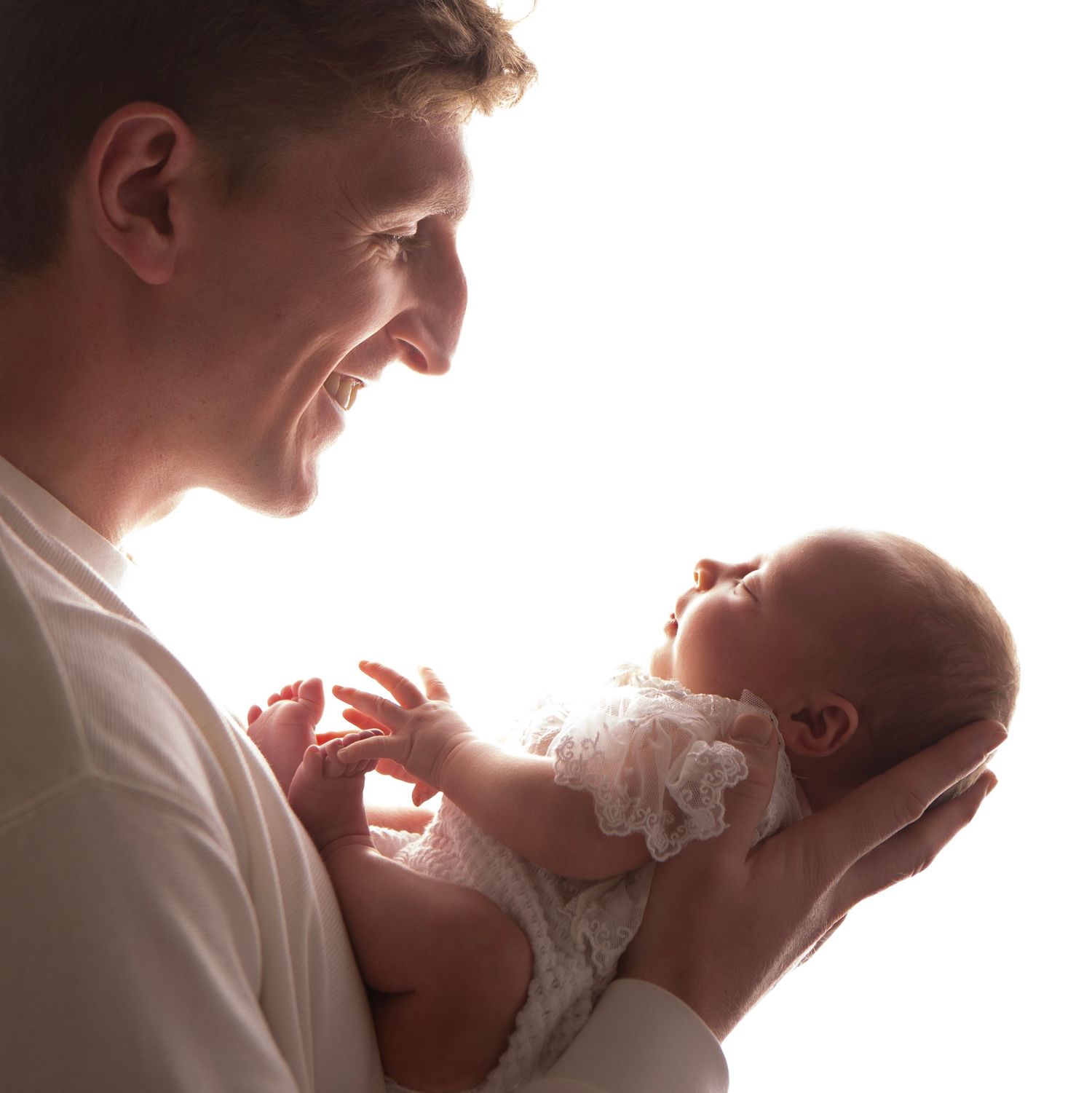 Smiling father holds newborn baby in white outfit, sharing a tender moment against bright white background.