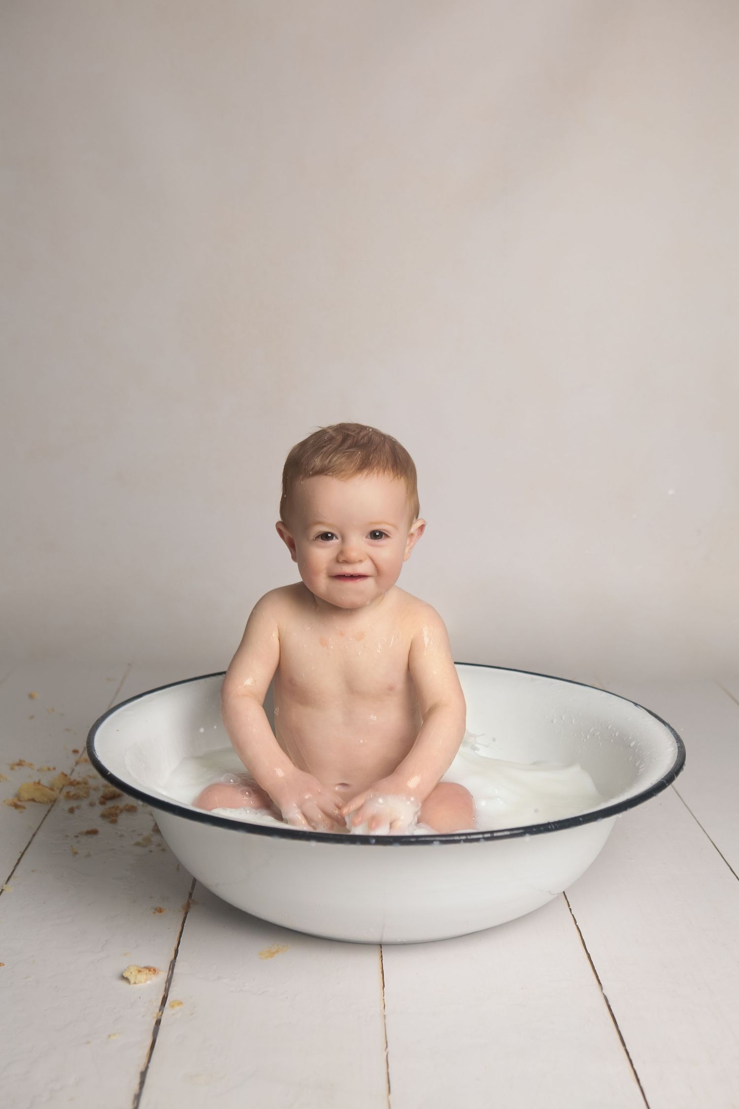 Smiling baby sitting in a large white enamel wash basin during a milk bath photoshoot on white wood floor.
