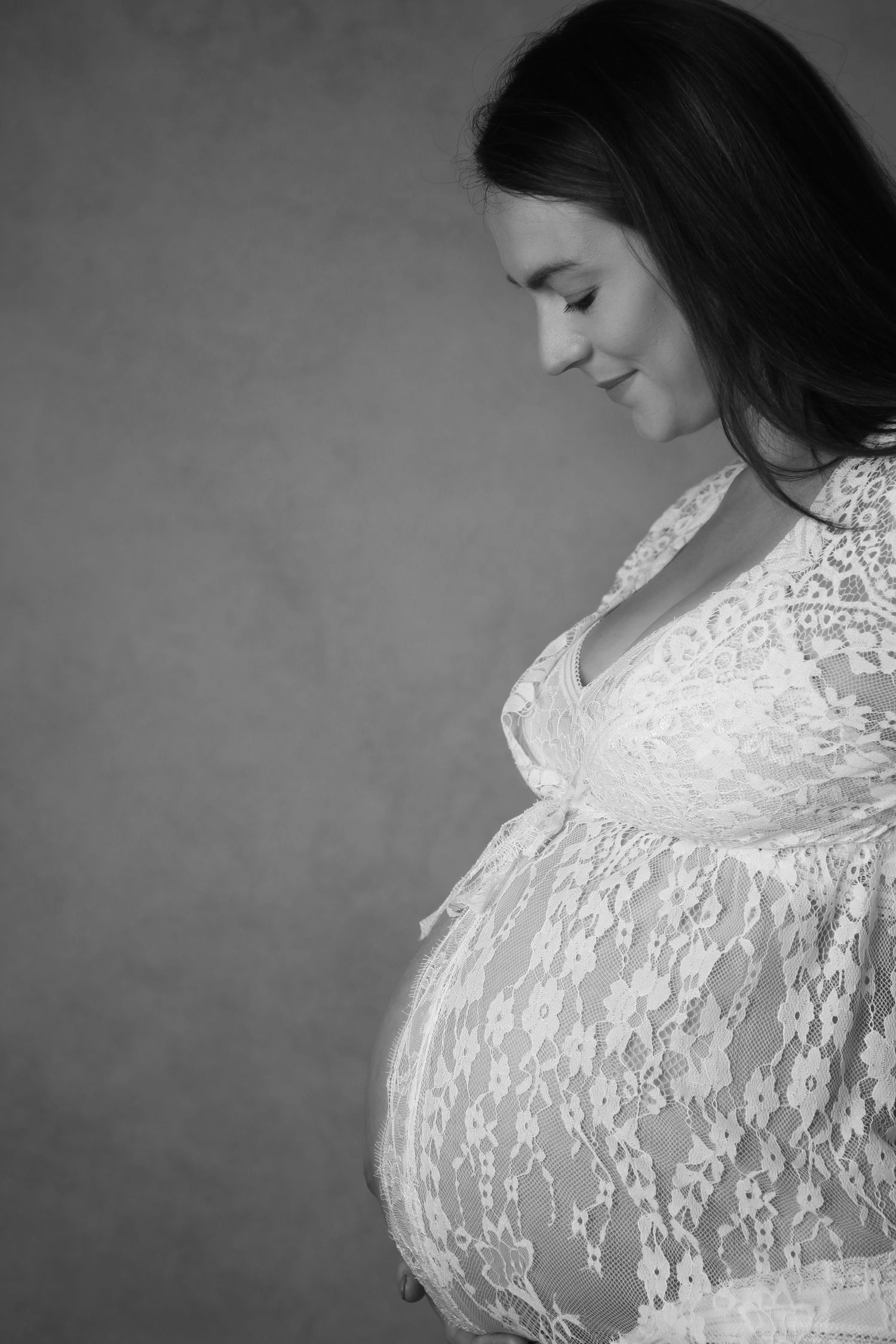 Black and white profile portrait of a smiling pregnant woman in lace dress cradling her baby bump.