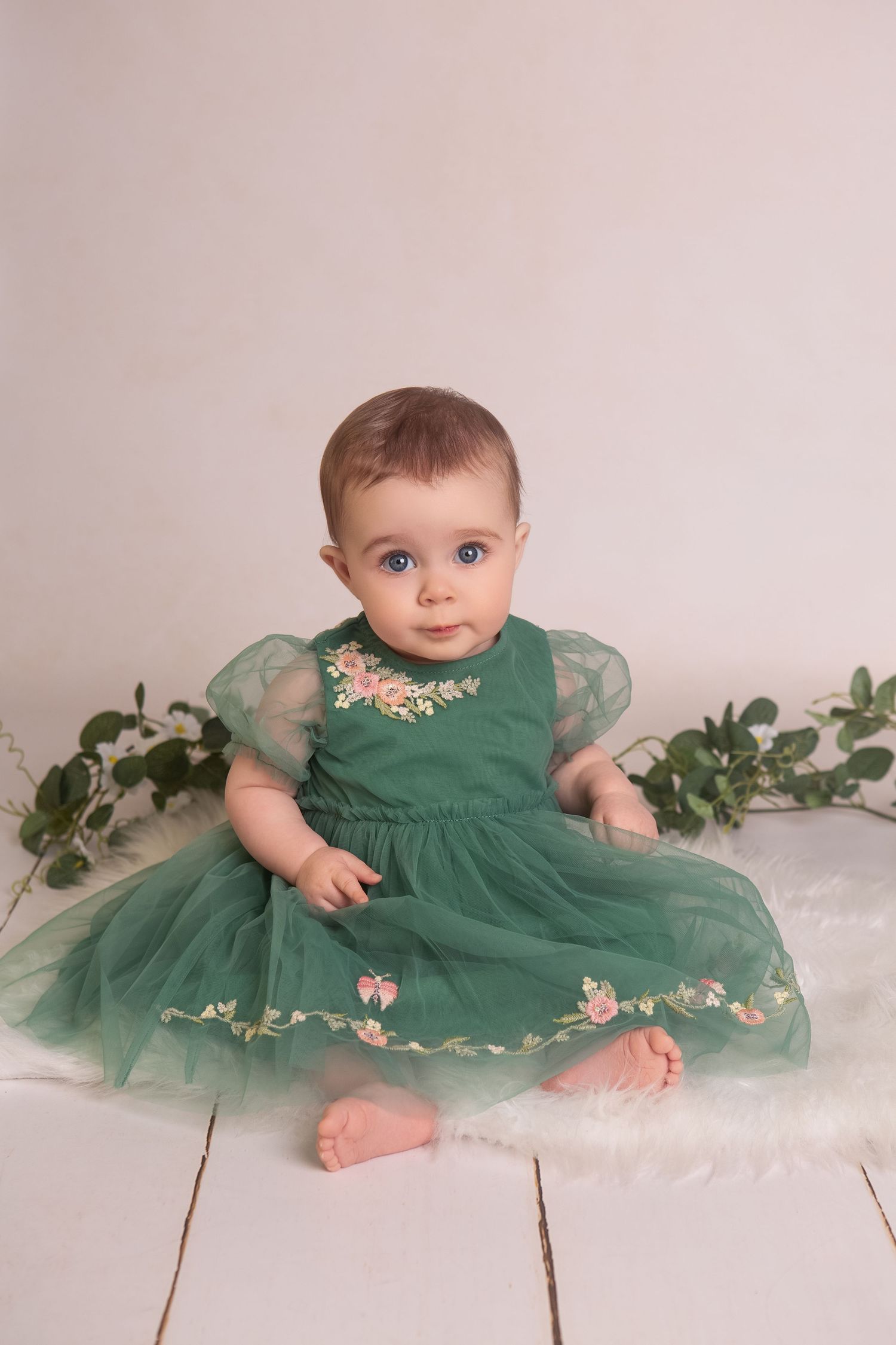 Baby girl in green floral tulle dress sitting on white fur rug with greenery backdrop.