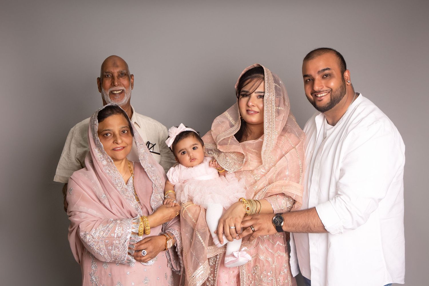 Multi-generational South Asian family in pink traditional attire posing together with a baby in a studio portrait.