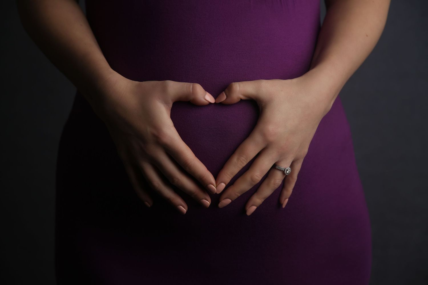 Pregnant woman in purple dress forming heart shape with hands on baby bump, maternity photography.