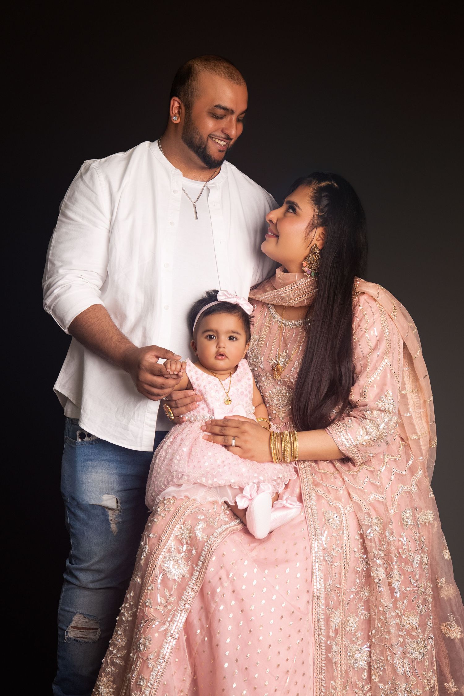 South Asian family portrait with man in white kurta, woman in pink lehenga, and baby girl in studio.