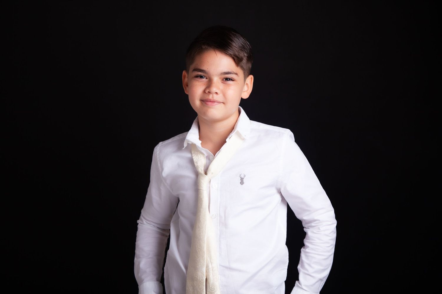Young boy in white dress shirt and tie smiling confidently against a black studio background.