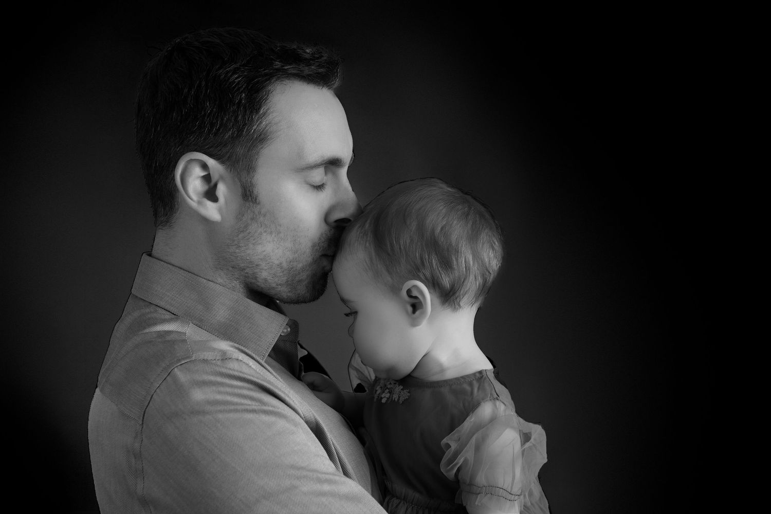 Black and white photo of a father tenderly kissing his baby on the forehead in a studio portrait.