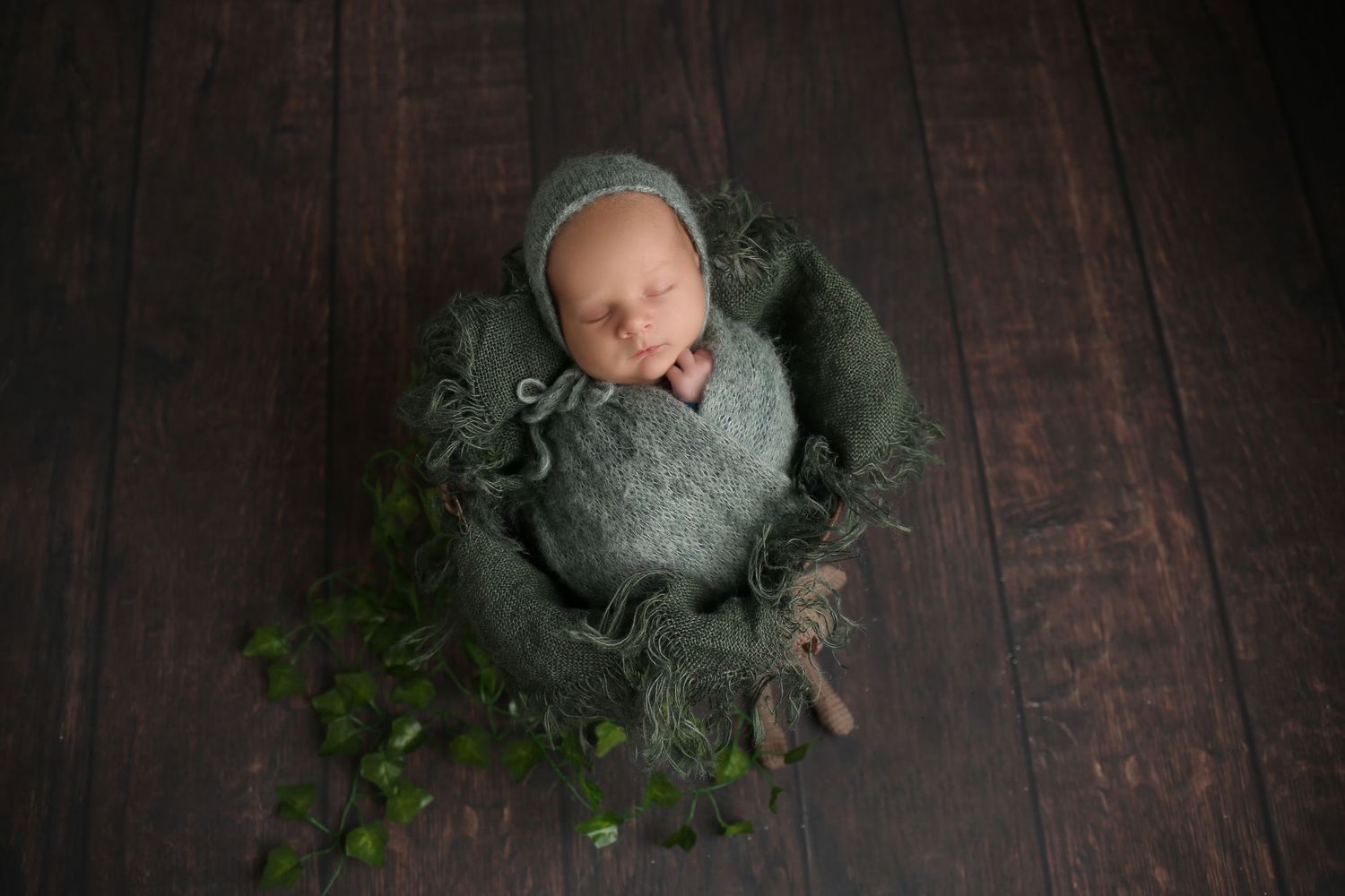 Newborn baby sleeping peacefully in a green knit bowl with ivy, wearing a sage bonnet on a dark wood floor.