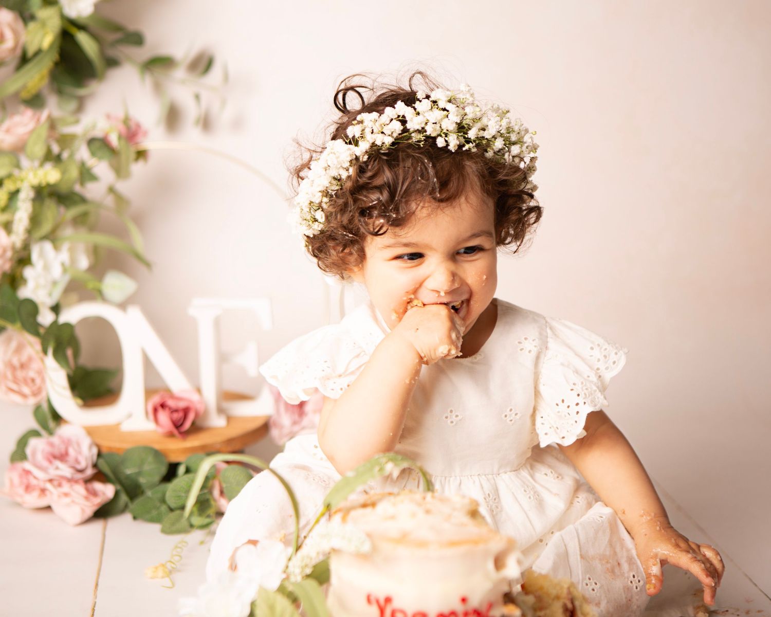 Joyful toddler in white lace dress and flower crown smashes her first birthday cake surrounded by pink roses.
