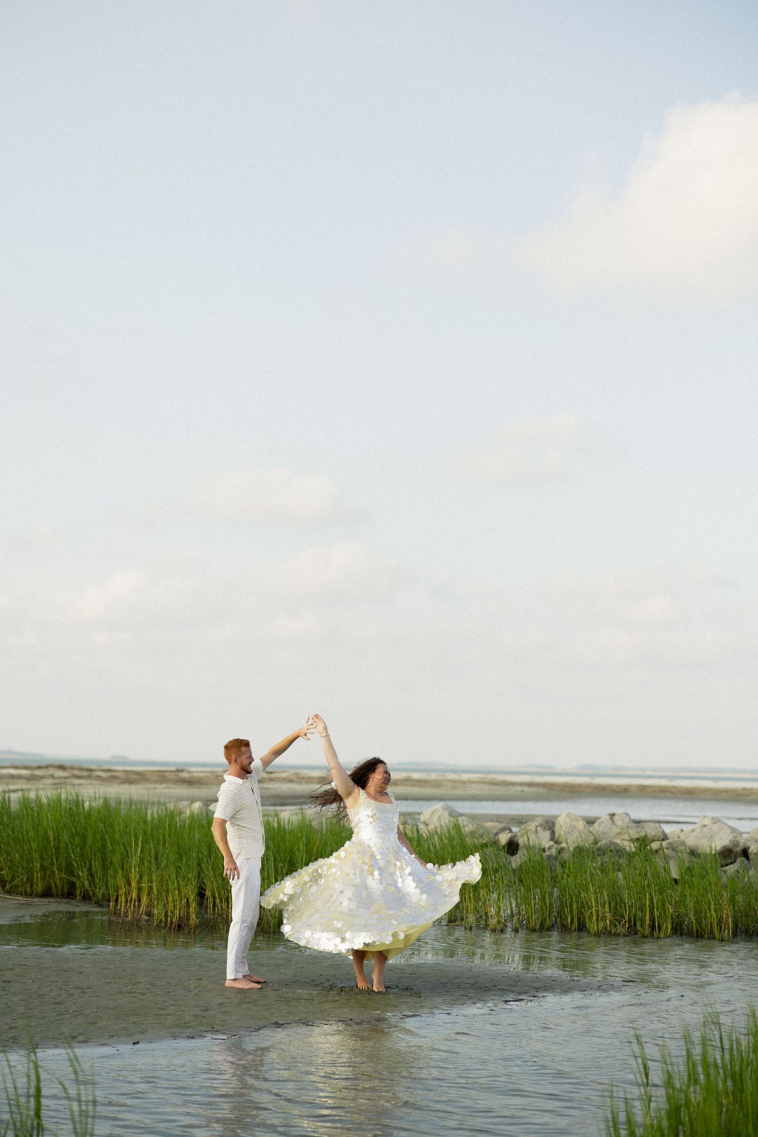 Couple in white dancing barefoot in shallow water near green marsh grass on a cloudy day.