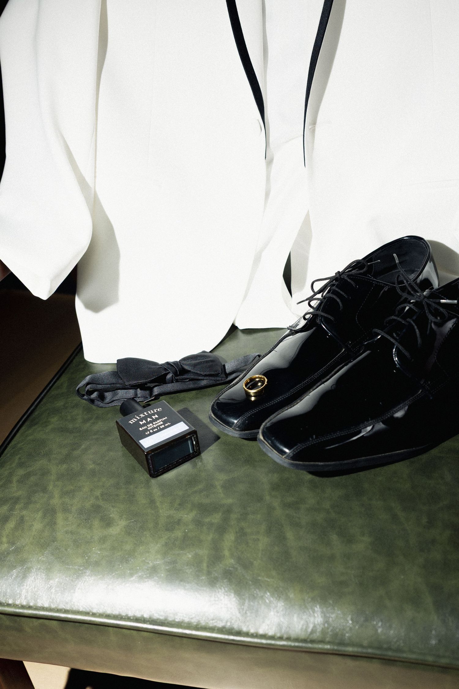 Black patent leather dress shoes, bow tie, cufflinks, and white tuxedo jacket arranged on a green leather bench.