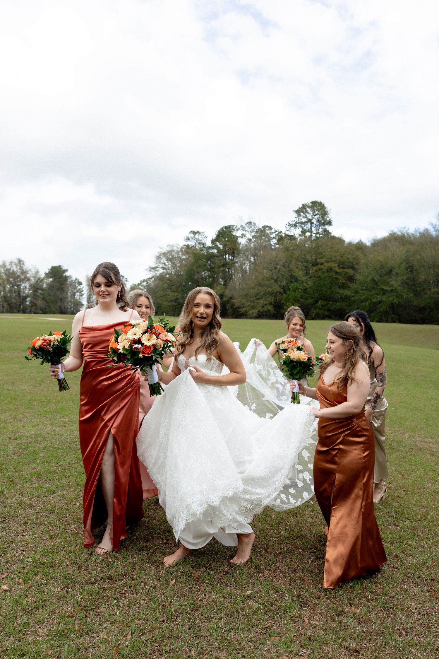 Bride in white gown walks with bridesmaids in rust orange satin dresses, holding bouquets on green lawn.