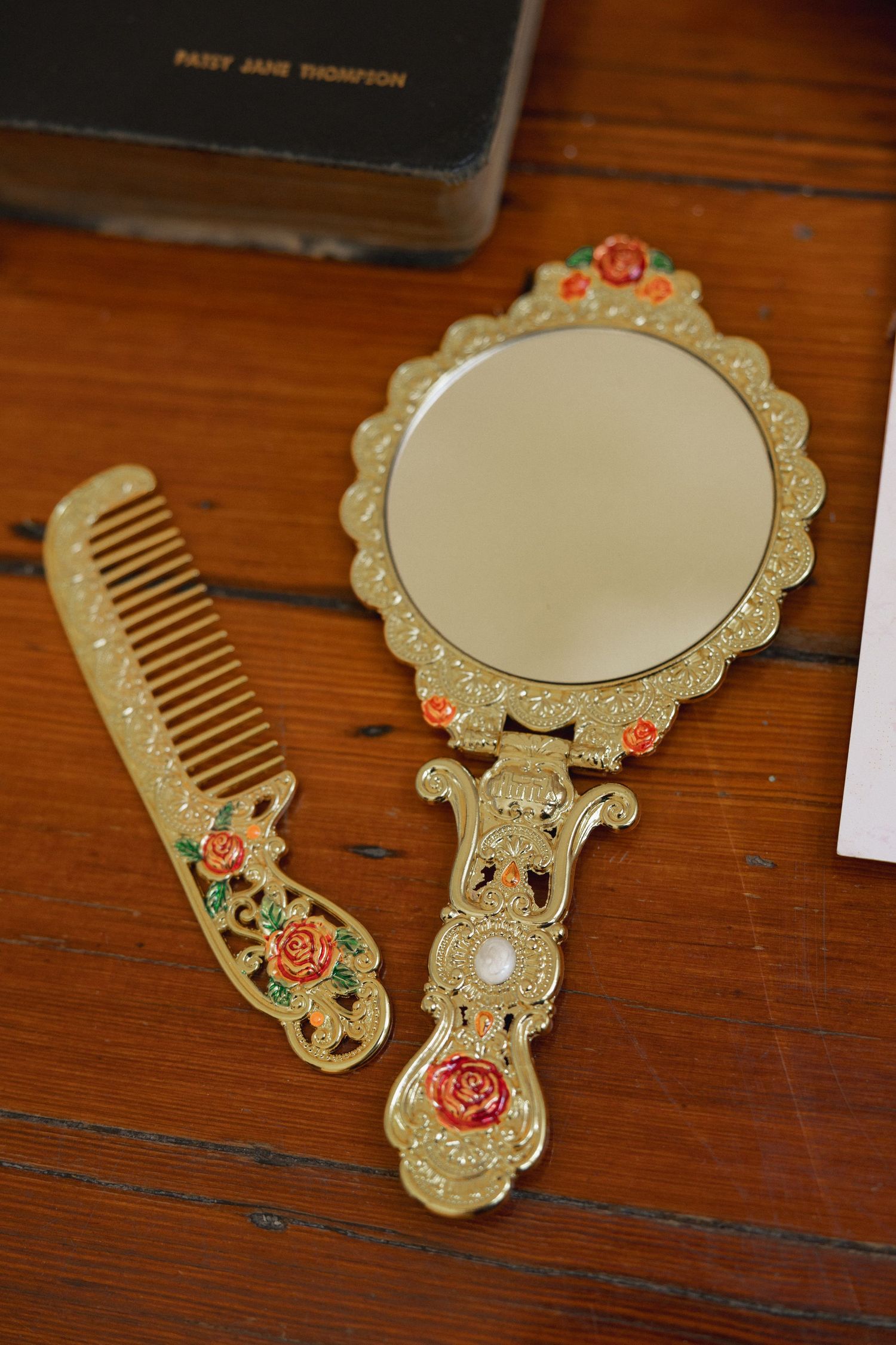 Ornate gold hand mirror and comb set with rose floral details on wooden surface beside vintage book.