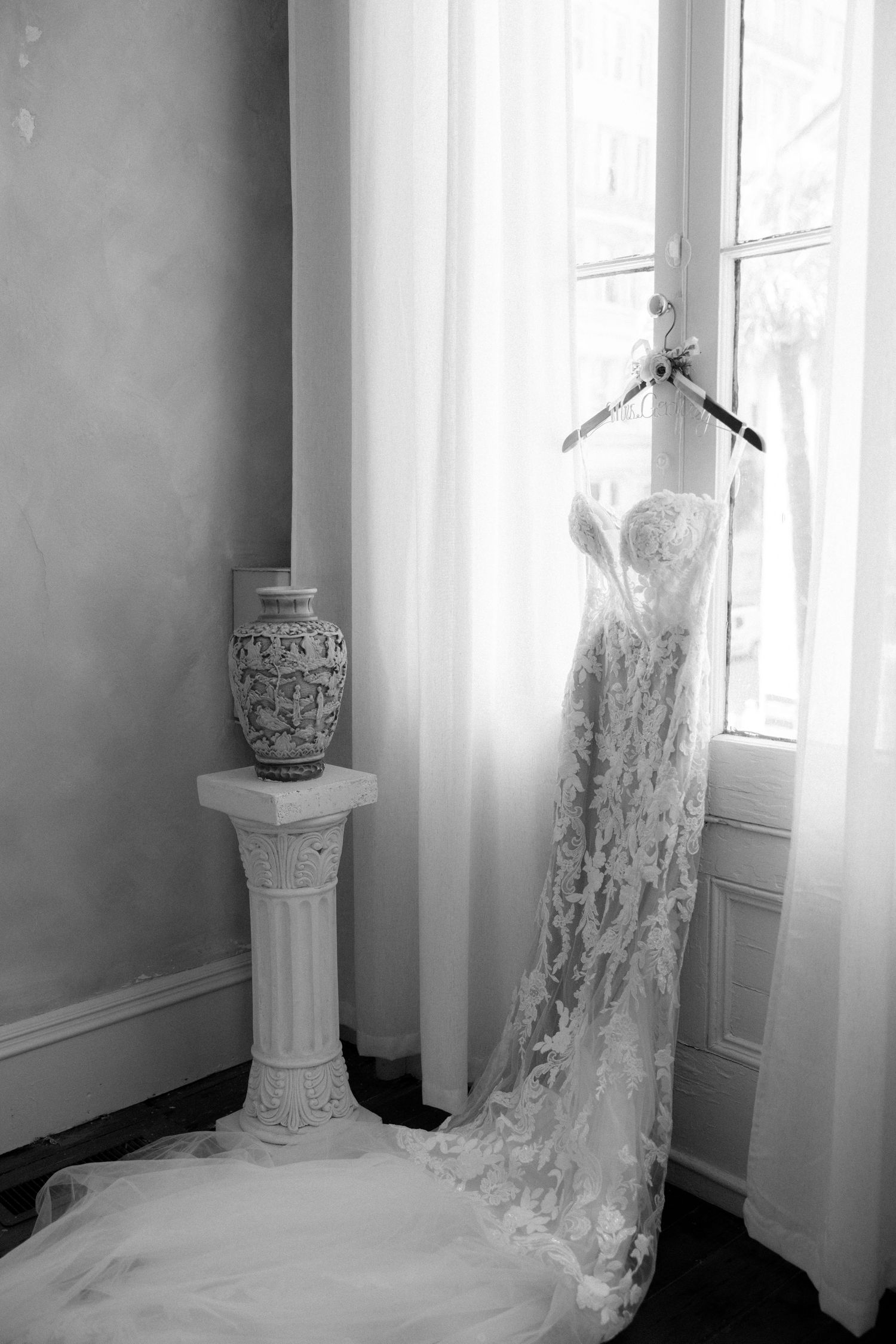 Elegant lace wedding dress hanging by a window with sheer curtains next to a decorative vase on a pedestal.