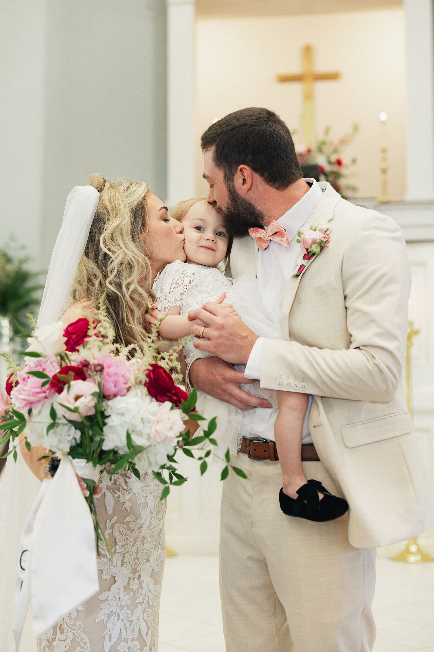Bride and groom kissing their baby at church wedding altar, groom in tan suit with pink bow tie, bride in lace gown holding florals.