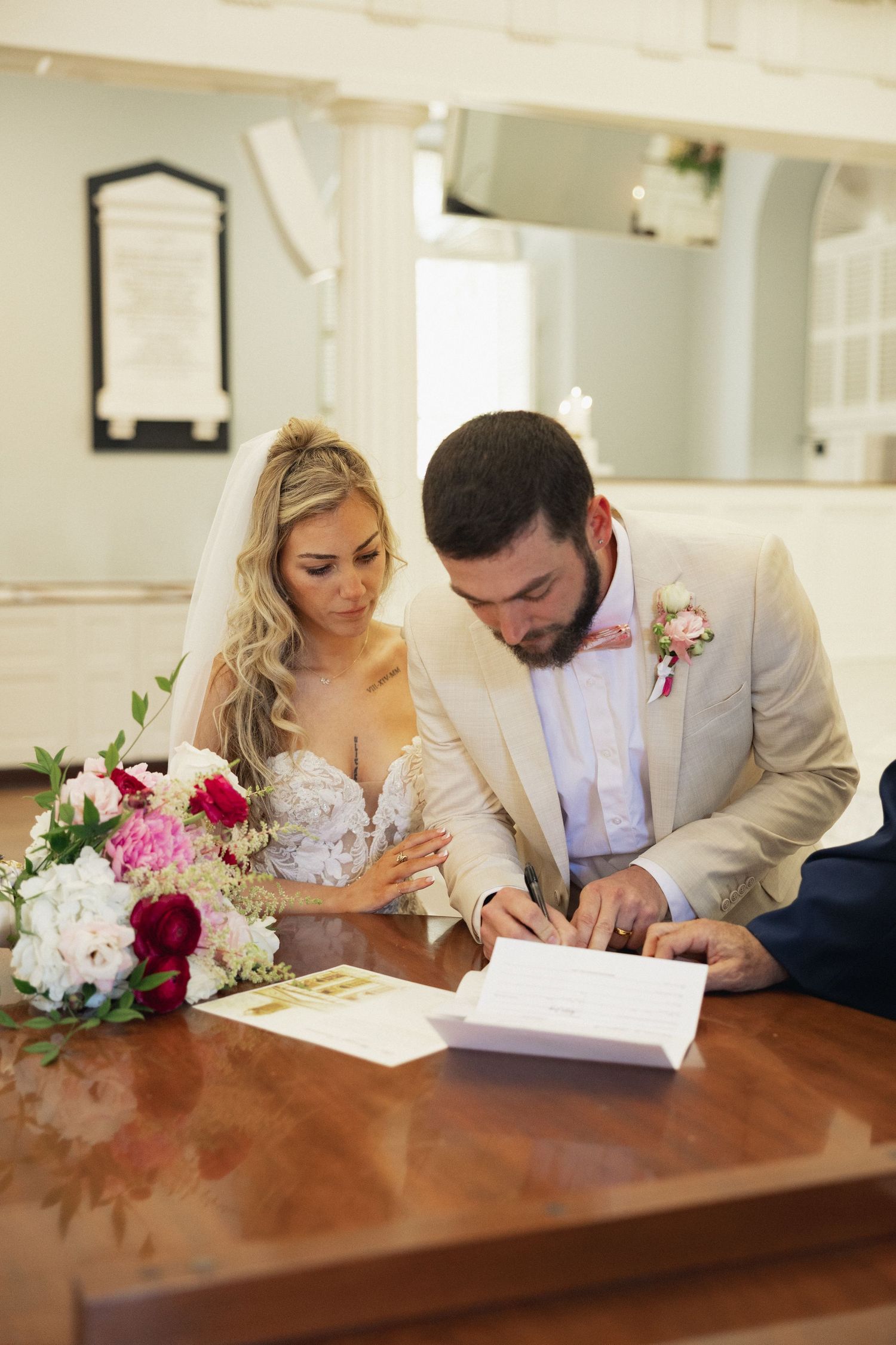 Bride and groom signing wedding documents at a wooden table with pink and white floral bouquet in a bright church.
